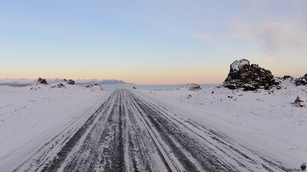 Blick auf einer verschneiten Straße, die gerade auf das Meer zuführt. Rechts sind dunkle Felsen am Straßenrand.