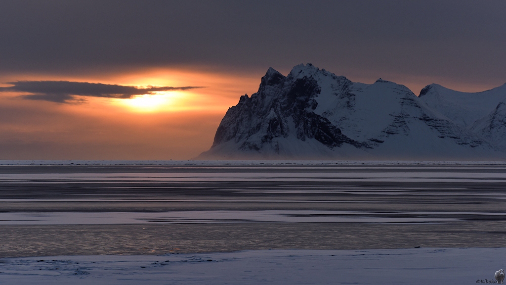Das Bild zeigt eine stimmungsvolle Gegenlichtaufnahme mit tiefstehender Sonne. Im Vordergrund ist eine teilweise zugefrorene Bucht mit Schneeflächen auf dem Eis. Felsige Berge bilden den Hintergrund.