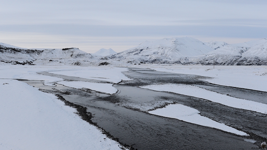 Das Bild zeigt einen verzweigten Fluss auf einer verschneiten Ebene. Im Hintergrund sind schneebedeckte Berge.