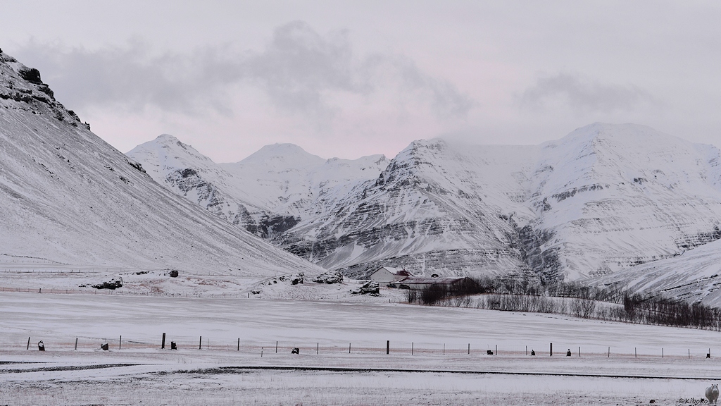 Das Bild zeigt einen Bauernhof auf einen Bergsattel. Im Hintergrund sind zwei Berge mit einem Taleinschnitt dazwischen. Im Vordergrund ist eine eingezäunte, verschneite Weidefläche.