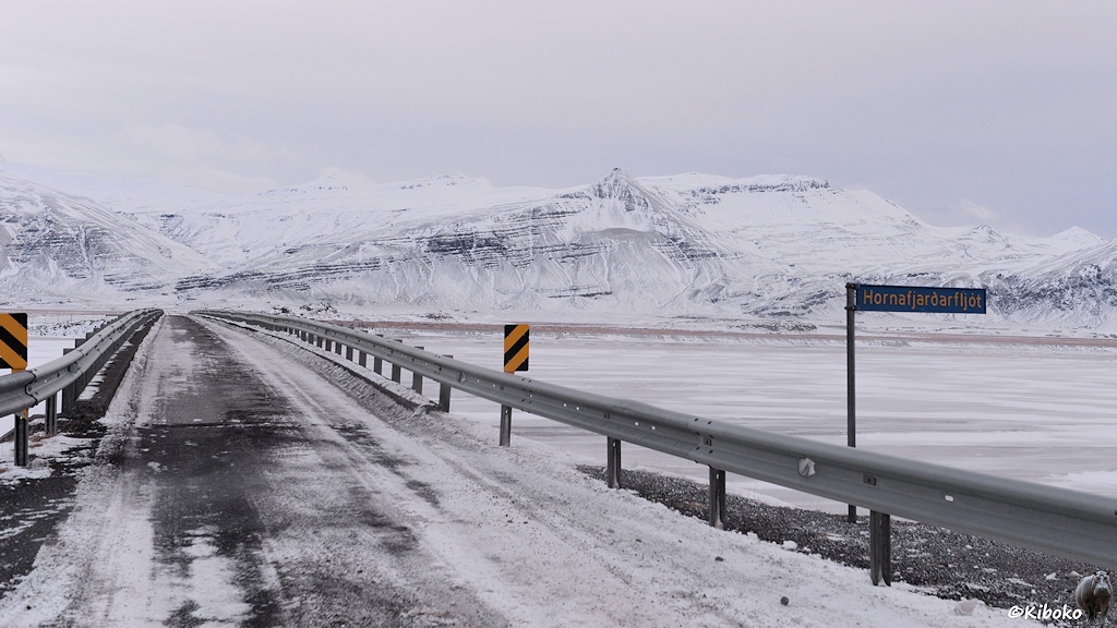 Das Bild zeigt eine einspurige Brücke über einen breiten vereisten Fluss. Auf einem blauen Schild mit gelber Schrift steht der Name des Flusses: Hornafjarðarfljót.
