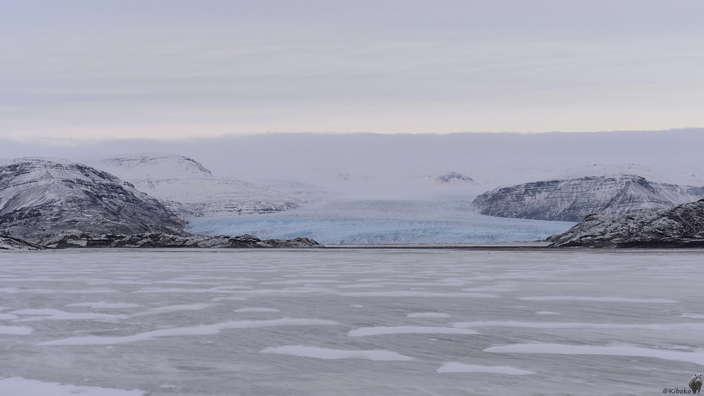 Das Bild zeigt eine vereiste Ebene, über der der Wind den Schnee treibt. Dahinter ist ein Gletscher aus hellblauen Eis, der durch Berge flankiert wird. Im Hintergrund sind Berge mit einem mächtigen, weißen Gletscher zu erkennen.