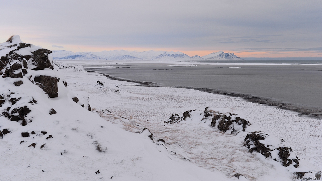 Das Bild zeigt eine verschneite Steilküste mit schwarzen Felsen von oben. Hinter einen schmalen schwarzen Strandstreifen ist der graue Atlantik. Im Hintergrund ist weit entfernt eine Bergkette. Die Spitzen der Berge liegen in einen pastellfarbenen Streifen in einer undefinierbaren Farbe zwischen gelb und rosa.