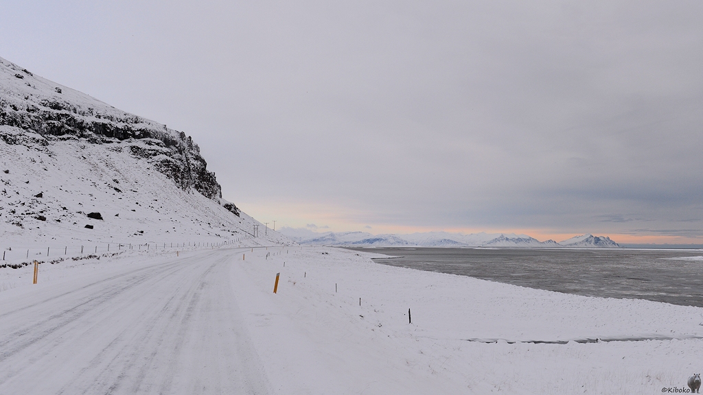 Das Bild zeigt eine schneebedeckte Straße vor einer Steilwand aus schwarzen Felsen. Am rechten Bildrand ist einer verschneiter Streifen und das graue Meer. Die Bergkette im Hintergrund ist die selbe, wie auf dem vorherigen Bild.