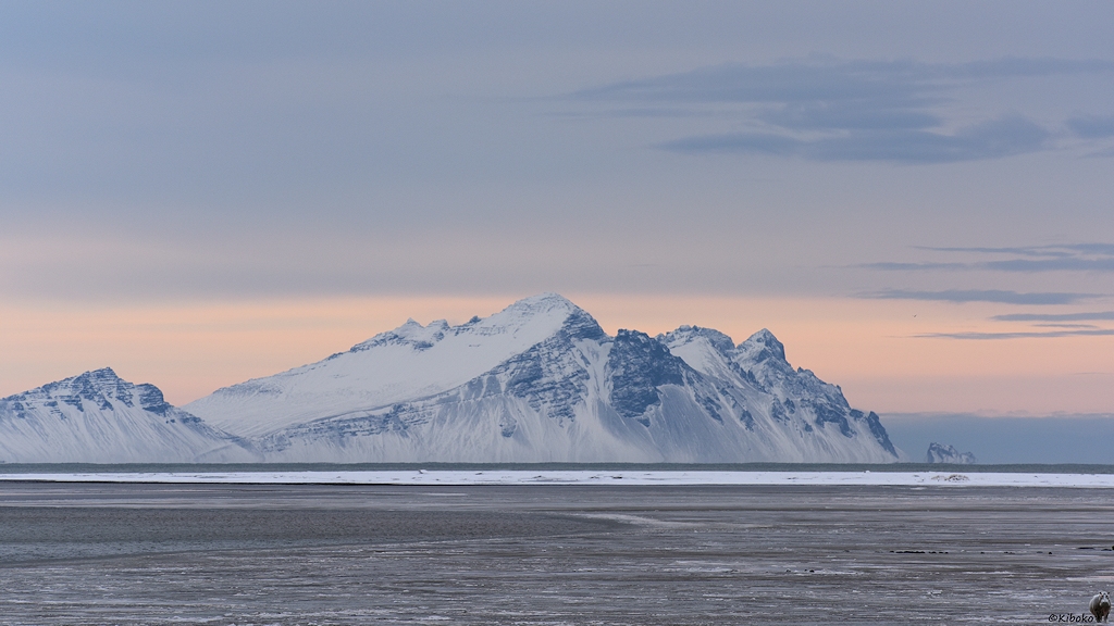 Das Bild zeigt einen verschneiten Berg an der Küste. Eine verschneite Sandbank trennt eine graubraune Lagune im Vordergrund vom grauen Meer im Hintergrund. Es ist eine Teleaufnahme von der Bergkette der vorhergehenden Bilder.