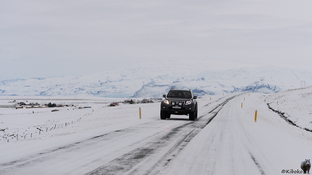Das Bild zeigt eine schneebedeckte Straße, die in das Bild führt. Ein dunkler Geländewagen mit viel Bodenfreiheit kommt auf der Straße entgegen. Links neben der Straße ist ein einer mittleren Entfernung eine Ortschaft. Im Hintergrund ist eine Bergkette, die im diffusen Licht verschwindet. Die Gipfel der Berge gehen übergangslos in den grauen Himmel über.