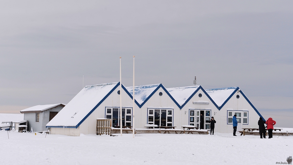 Das Bild zeigt ein weißes, einstöckiges Holzhaus im Schnee. Das Haus hat ein vierzackigen Giebel, als wenn vier kleine Häuschen aneinandergereiht sind. Das Dach ist mit Schnee bedeckt. Vor dem Haus stehe Holztische und Holzstühle, die ebenfalls mit Schnee bedeckt sind.