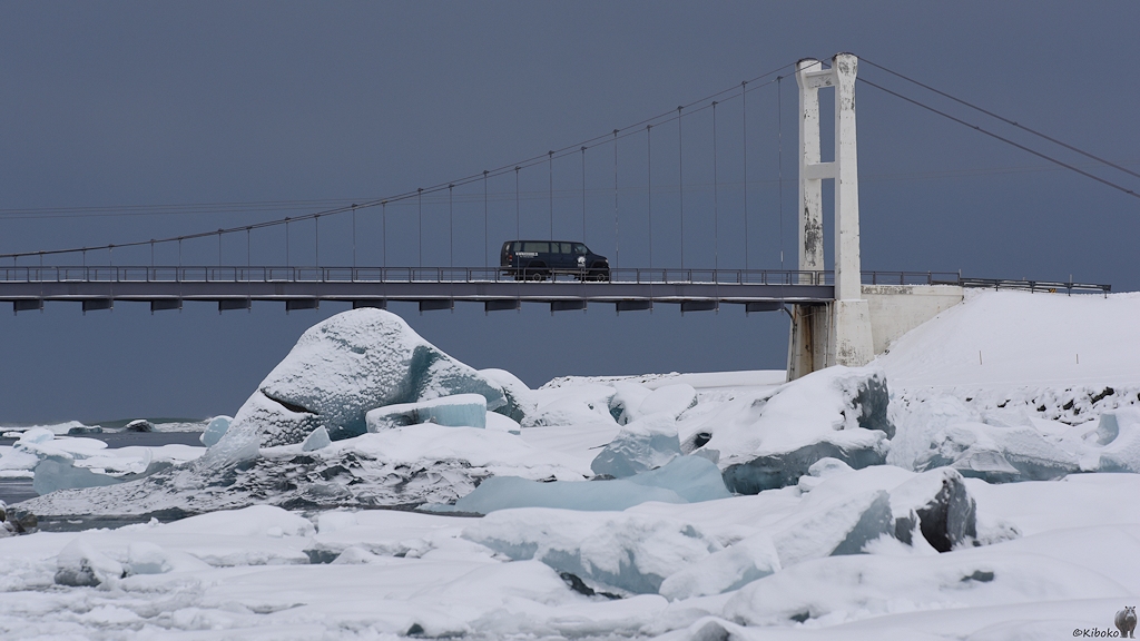 Das Bild zeigt ein Teil der Hängebrücke mit dem rechten Pylonen mit dunkelgrauem Himmel im Hintergrund. Über die Brücke fährt ein dunkelgrauer Kleinbus. Unter der Brücke sind verkeilte Eisberge aus hellblauen Gletschereis, die mit Neuschnee überzuckert sind.