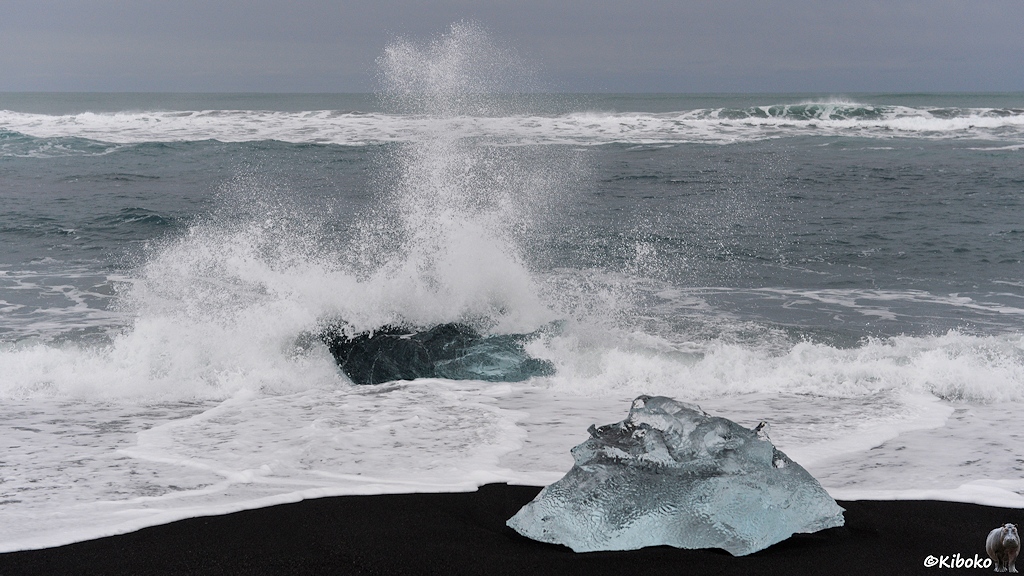Das Bild zeigt einen Eisblock in der Brandung. Das Wasser spitzt hoch. Die Welle läuft weiter entgegen einen zweiten hellblauen Eisblock am schwarzen Strand.