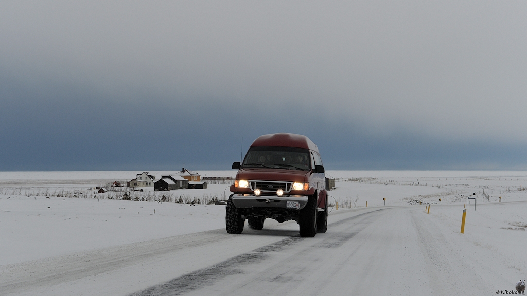 Das Bild zeigt einen höhergelegten, roten Kleinbus der auf einer verschneiten Straße entgegenkommt. Der Kleinbus hat dicke Reifen mit Spikes. Im Hintergrund sind ein paar Häuser auf einer verschneiten Ebene. Der Himmel ist dunkelgrau.