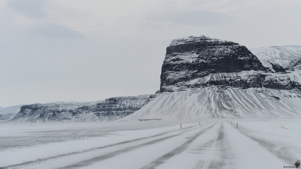 Das Bild zeigt eine verschneite Straße die auf einen mächtigen schwarzen Felsen führt. Vor dem Felsen biegt die Straße nach links, führt über eine Brücke an einer Felswand entlang. Links ist eine verschneite Ebene. Der Wind weht Schnee über die Straße und die Ebene.