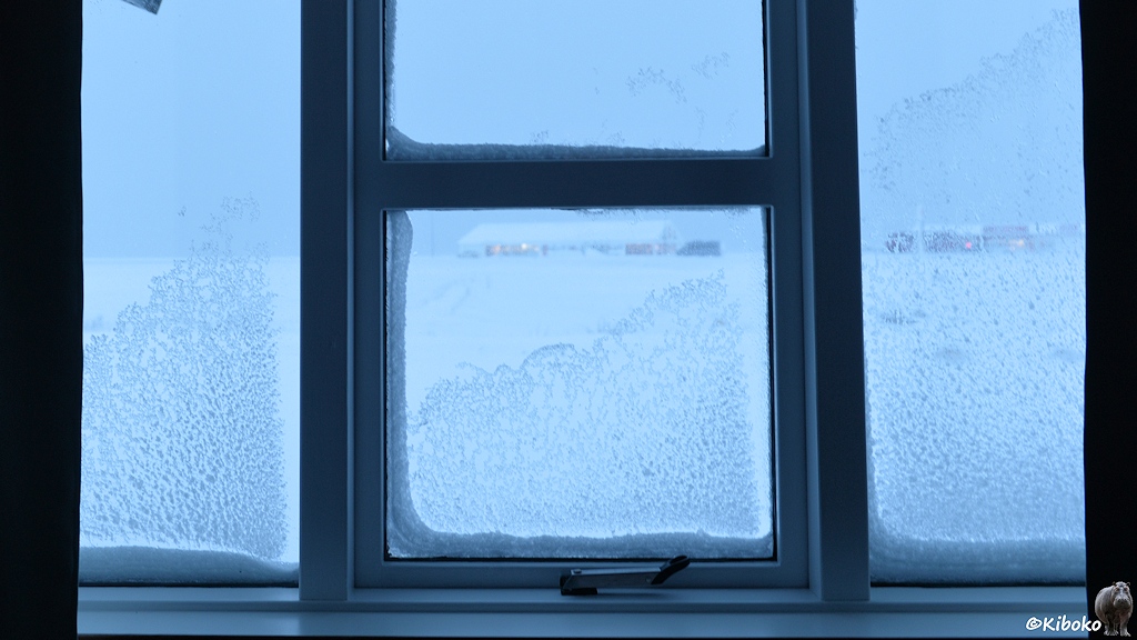 Das Bild zeigt einen Blick durch das Fenster. Der Schnee klebt am Fensterglas. Die beleuchteten Nachbarhäuser sind während der einsetzenden Dämmerung kaum zu sehen.