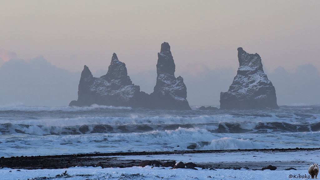 Das Bild zeigt drei Felsnadeln im aufgewühlten Meer. Die Felsnadeln haben eine dünne Schneeschicht. Der Horizont ist trübe und grau.