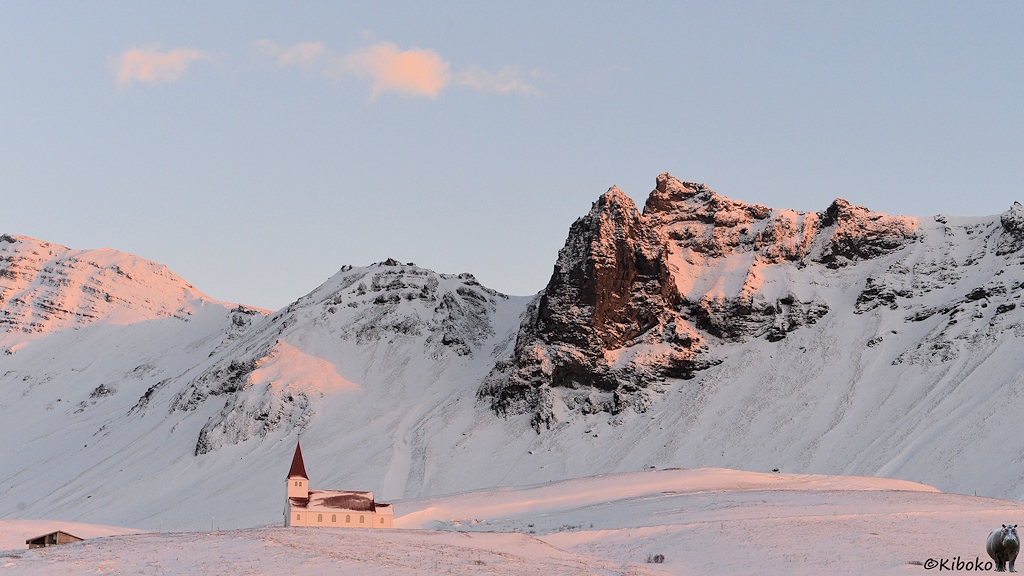 Das Bild zeigt eine kleine weiße Kirche vor einer verschneiten Bergkulisse. Die Kirche wird von der tiefstehenden Morgensonne angestrahlt. Dir Kirche hat ein rotes Dach, vier Seitenfenster und einen viereckigen Turm über dem Eingang.