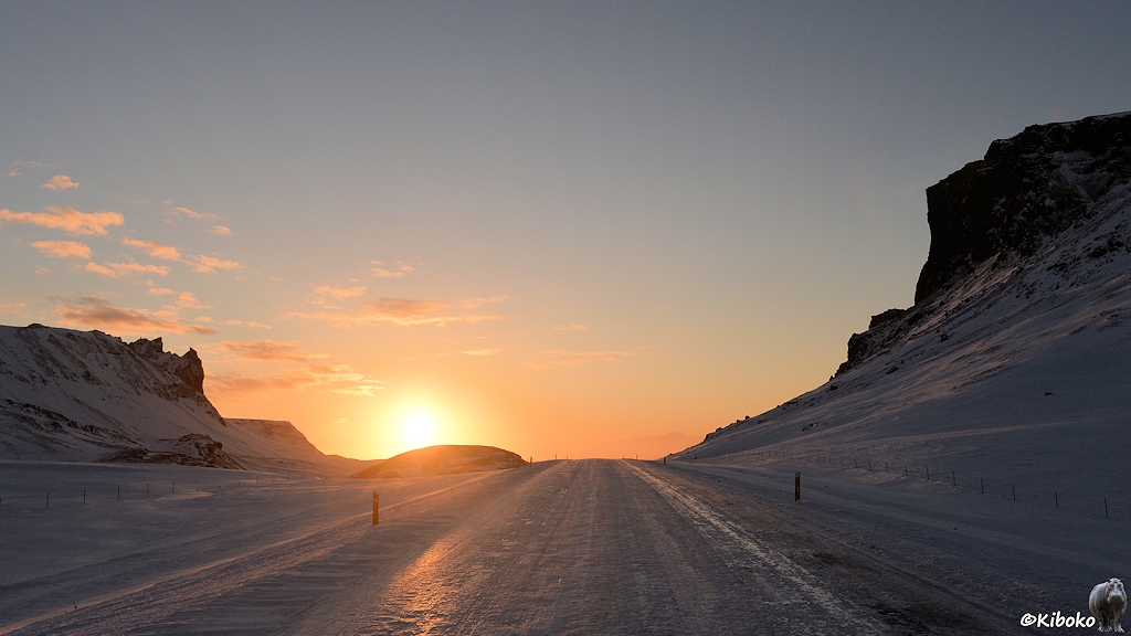 Das Bild zeigt eine vereiste Straße die geradlinig über einen Pass führt. Auf beiden Seiten sind verschneite Felswände. Die Sonne scheint gerade so über die Passhöhe und taucht Himmel und Straße in oranges Licht.
