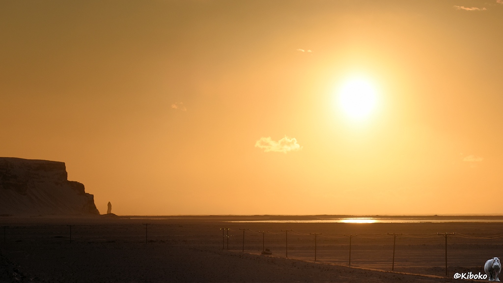 Das Bild zeigt einen schwarzen Strand mit einer Lagune davor. Die Sonne steht hoch über den Strand. Der Himmel leuchtet orange. Am linken Bildrand ist ein steil abfallendes Kap mit einer Steinsäule im Meer.