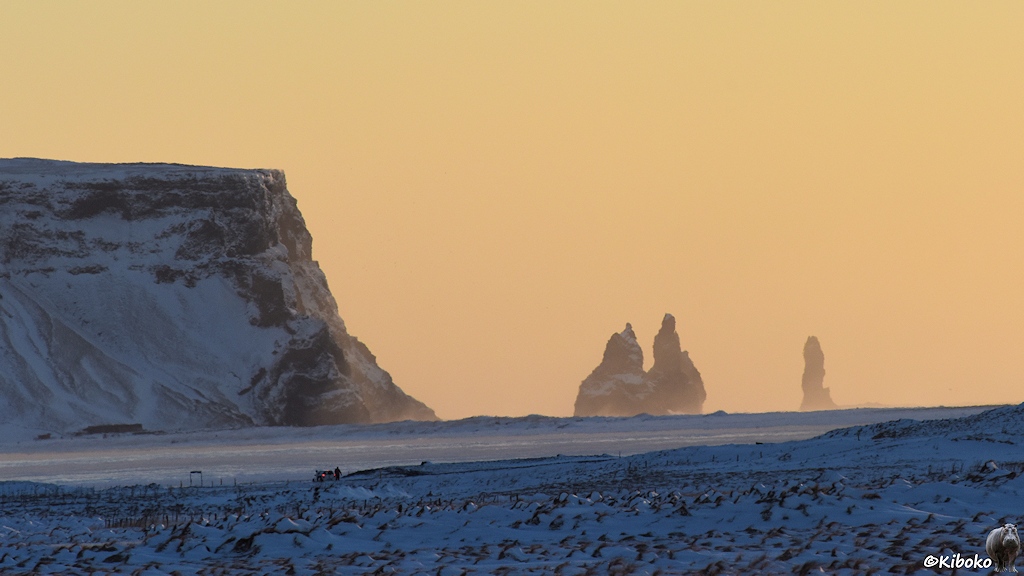 Das Bild zeigt eine verschneite Landschaft mit einem felsigen Kap auf der Linken Seite und Steinsäulen in der Mitte und der linken Seite. Der Himmel ist blass gelborange. Auf einer nicht einzusehenden Straße steht ein weißer Kleinwagen.