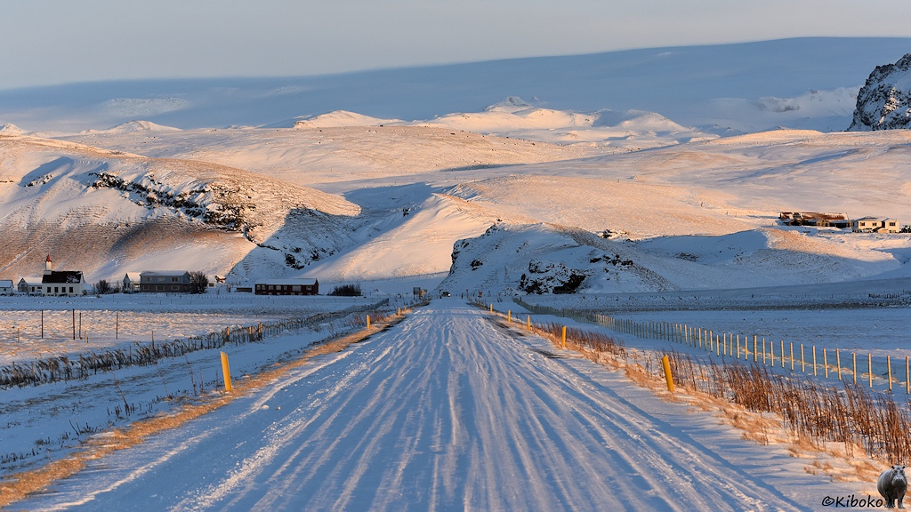 Das Bild zeigt eine schneebedeckte Straße, die gerad ein das Bild führt. Im Hintergrund ist eine Bergkette und eine Ortschaft im Tal. Die Sonne taucht die Winterlandschaft von hinten in ein warmes Licht.