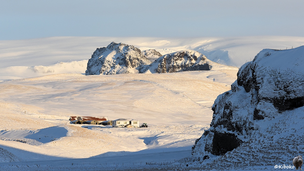 Das Bild zeigt einen Bauernhof in einer weiten Schneelandschaft. Im rechten Vordergrund und im Hintergrund sind große dunkle Felsen.
