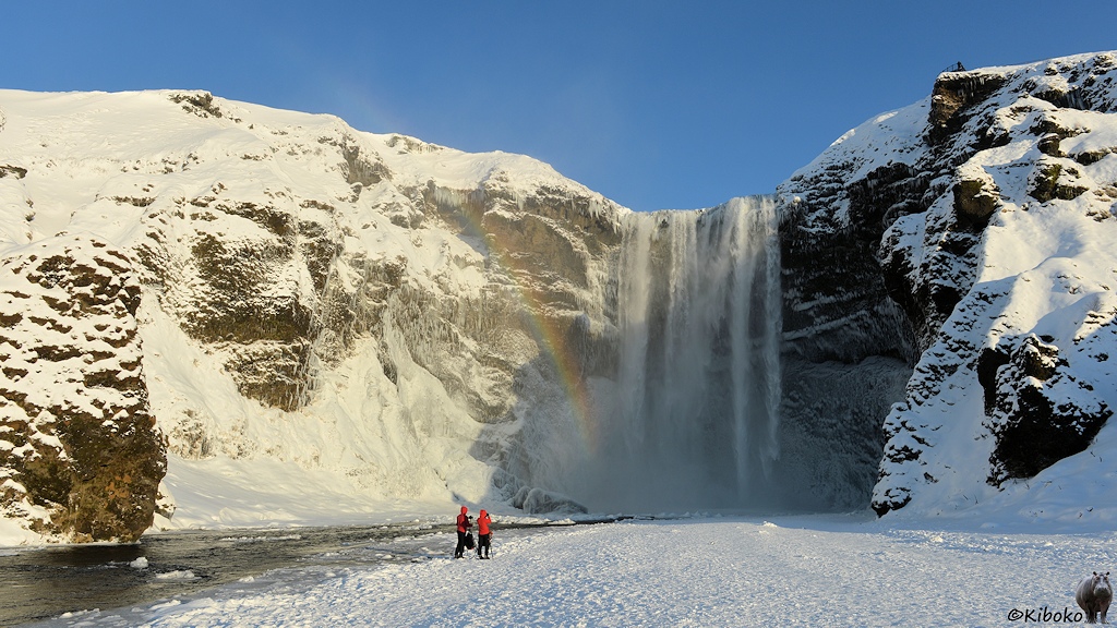 Das Bild zeigt einen Wasserfall, der eine 80m hohe verschneite Felswand herunterstürzt. Ein Regenbogen steigt vor dem Wasserfall auf. Zwei Fotografen stehen an einem Fluss vor dem Wasserfall.