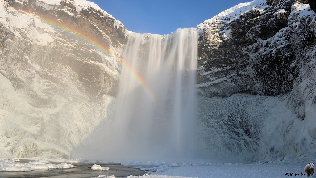 Das Bild zeigt einen Wasserfall im Sonnenlicht. Ein Regenbogen ist vor dem Wasserfall. Durch eine längere Belichtungszeit wirkt der Wasserfall, wie ein Vorhang. Auf beiden Seiten sind dunkle Felsen. Die Vorsprünge sind mit Schnee und Eiszapfen bedeckt.