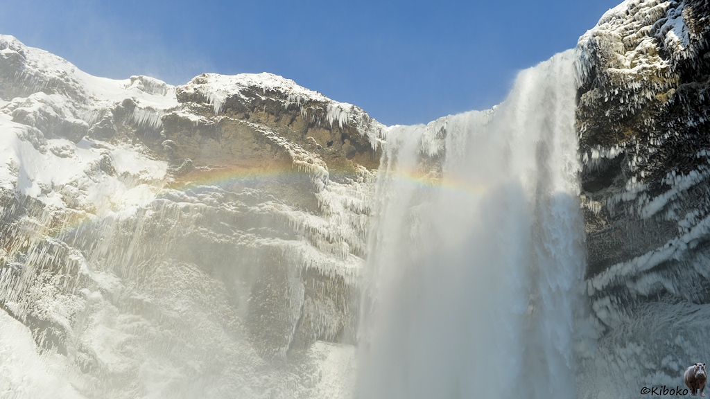 Das Bild zeigt den oberen Teil eines Wasserfalls der sich breit in Tiefe stürzt. Ein Regenbogen verläuft im flachen Bogen quer vor dem Wasserfall und die benachbarte4n dunklen Felswände. Schnee und Eiszapfen verzieren die Felswände.