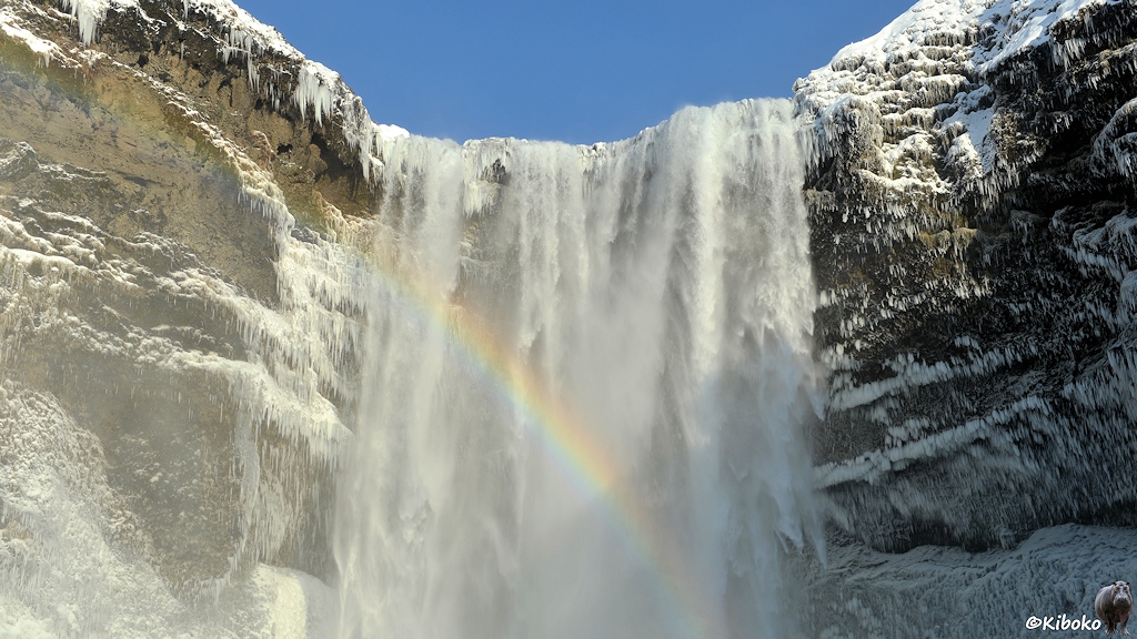 Das Bild zeigt den oberen Teil eines Wasserfalls direkt von vorn. Ein Regenbogen steigt von rechts unten nach links oben vor dem Wasserfall empor.