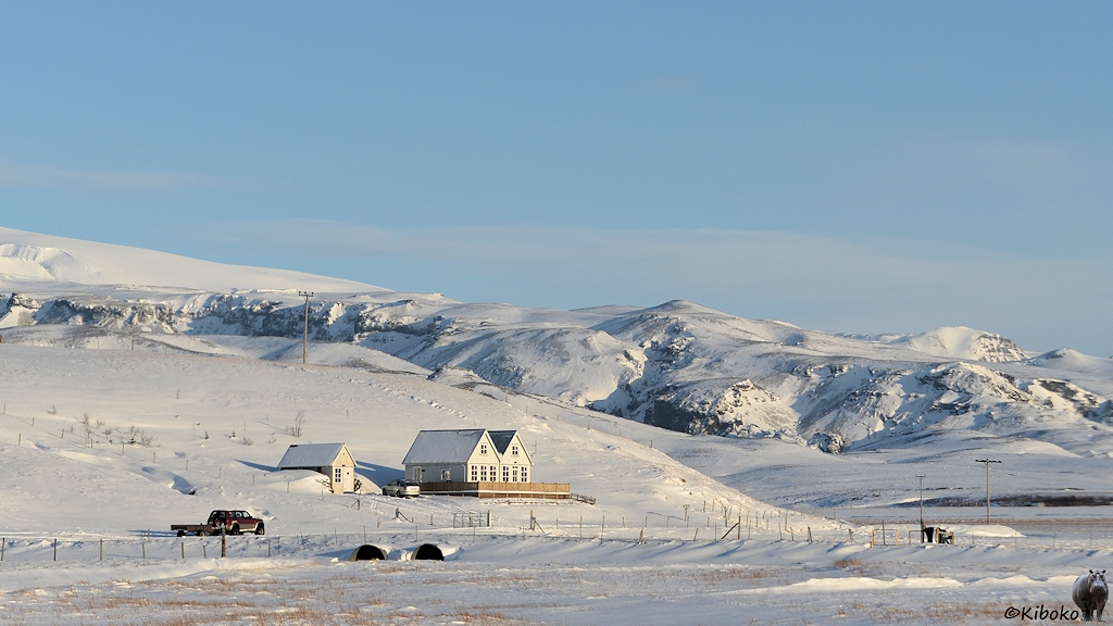 Das Bild zeigt eine weite Winterlandschaft mit Bergen im Hintergrund. An einem Fuß des Berges ist ein weißes Doppelhaus mit großer eingezäunter Veranda. Davor steht ein Geländewagen mit Anhänger.