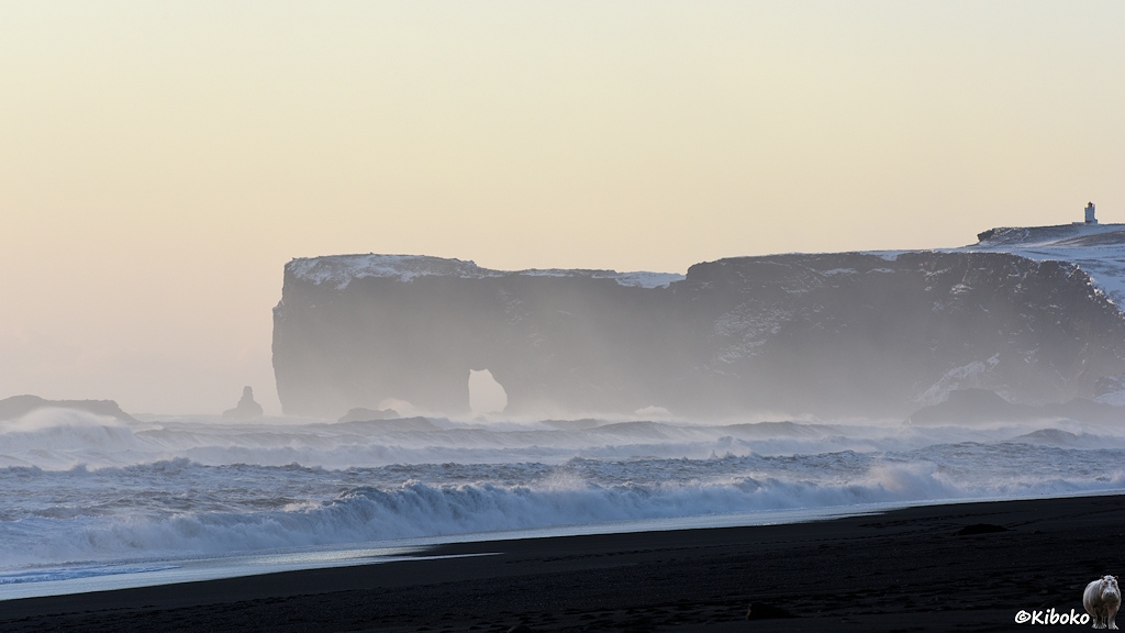 Das Bild zeigt ein Kap mit einem Felsentor an der Spitze. Durch die große Entfernung und der Gischt der Wellen ist der scharze Fels grau. Im Vordergrund ist der Atlantik mit Wellen, die auf einen schwarzen Sandstrand treffen.