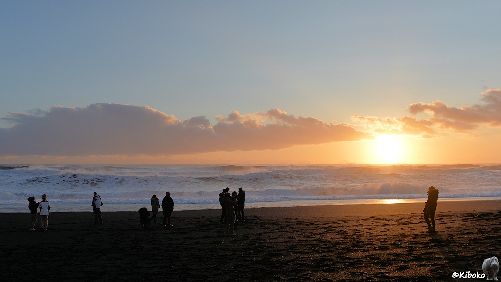 Das Bild zeigt Menschen auf einem schwarzen Sandstrand bei Sonnenuntergang. Das aufgewühlte Meer schäumt weiß.