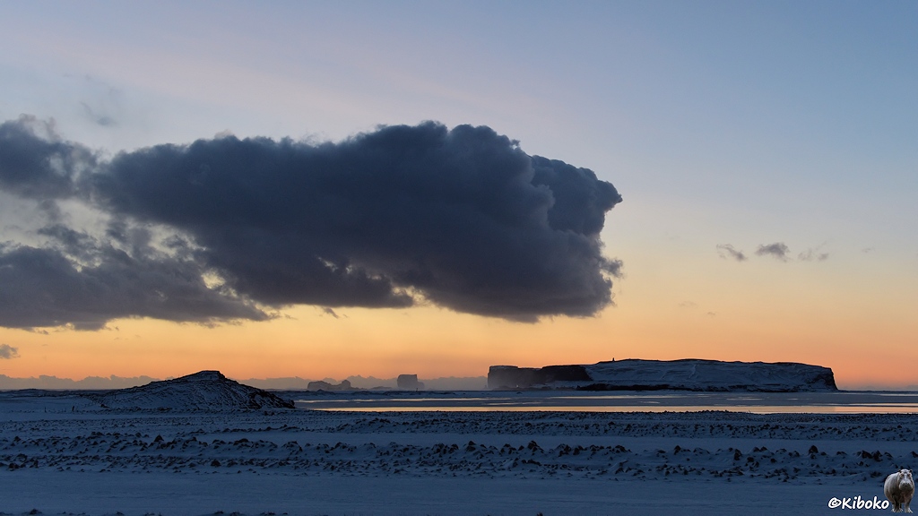 Das Bild zeigt eine Landschaftsaufnahme nach Sonnenuntergang. Zentral im Bild ist ein großer flacher Fels mit dem Kap Dyrhóley. Darüber schwebt eine große schwarze Wolke. Am Horizont ist ein oranger Streifen. Der Himmel darüber und der Schnee im Vordergrund sind bläulich.