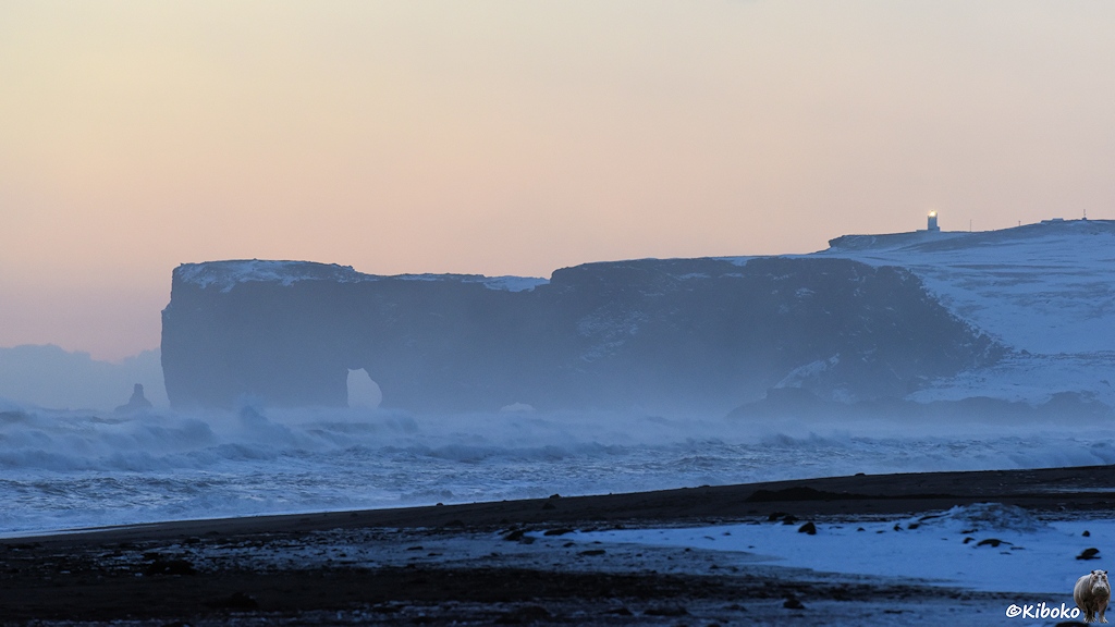 Das Bild zeigt Teleaufnahme von einem Kap mit Felsentor in der Dämmerung. Darauf steht ein Leuchtturm. Das Licht des Leuchtturms ist an. Im Vordergrund ist ein schwarzer Strand und aufgewühltes Meer.