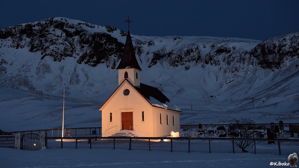 Das Bild zeigt eine kleine weiße Kirche, die angestrahlt wird. Sie hat eine braune Tür und darüber ein kleines viereckiges Türmchen. Die Kirche ist eingezäunt. Hinter der Kirche ist ein schneebedecktes Bergmassiv in der Dämmerung.