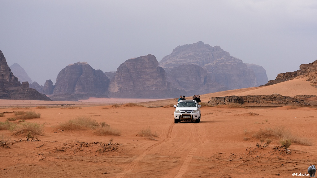 Weißer Geländewagen in ornageroter Wüste vor Bergkulisse mit wolkenverhangenen Himmel