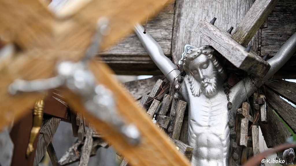 Das Bild zeigt den Oberkörper eines silbernen Kruzifix an dessen Arme viele kleine Holzkreuze hängen. Im Vordergrund ist auf der linken Seite ein weiterer Kruzifix unscharf dargestellt.