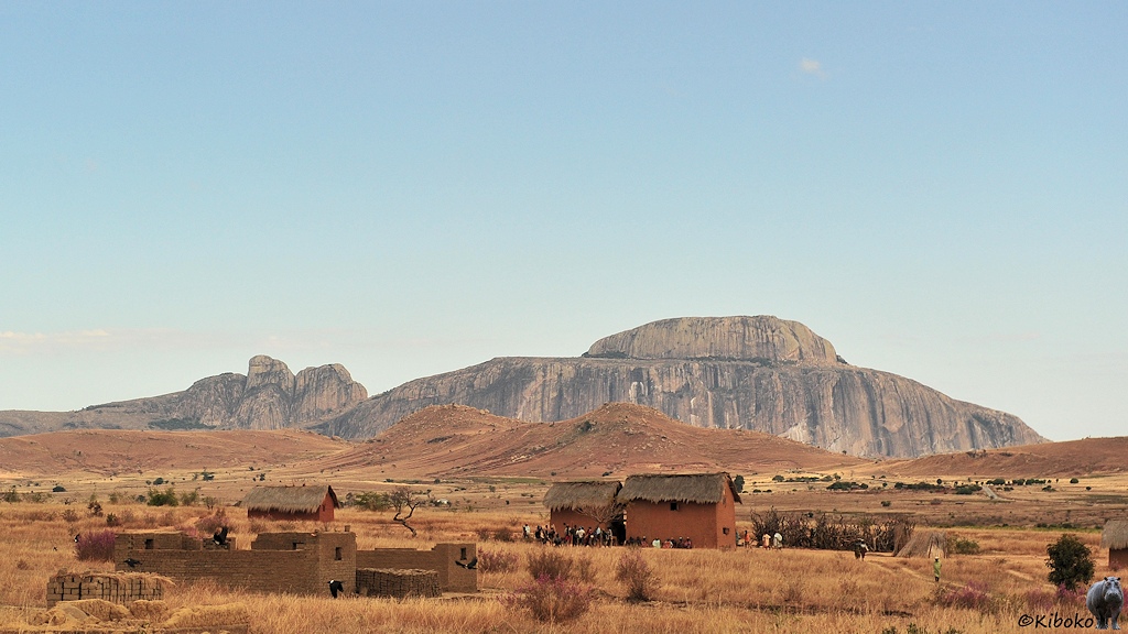 Das Bild zeigt einen breiten Felsenberg mit einen Felsenaufsatz in der Mitte. Er hat die Form eines Hutes. davor sind ein paar Hügel. Im Vordergrund ist ein Dorf aus kleinen rotbraunen Häusern mit Strohdach in einer Ebene aus trockenem Gras.
