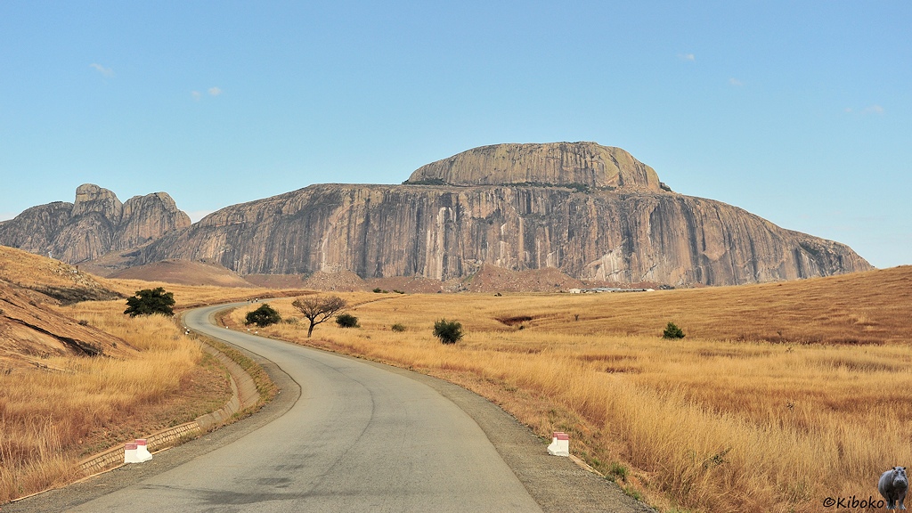 Das Bild zeigt eine Straße die in einer weiten S-Kurve auf einen Berg aus einem großen Felsen in Hutform führt. Sonst ist die Landschaft mit trockenem Gras bedeckt.