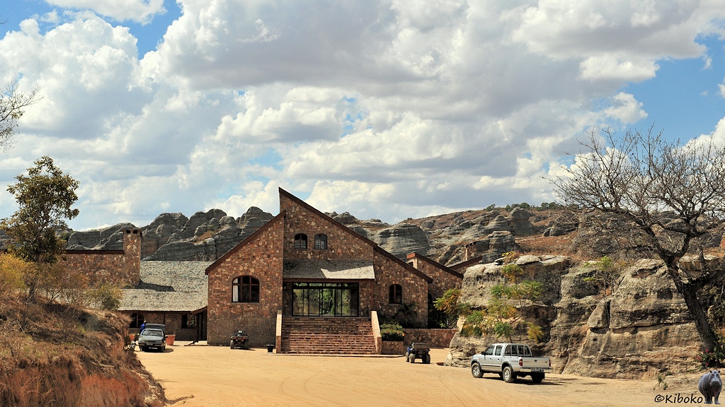 Das Bild zeigt ein Gebäude aus braunen Naturstein mit asymmetrischen Giebel und breiter Treppe eingebettet in einer felslandschaft. Davor ist ein sandiger Parkplatz mit mehreren Fahrzeugen.