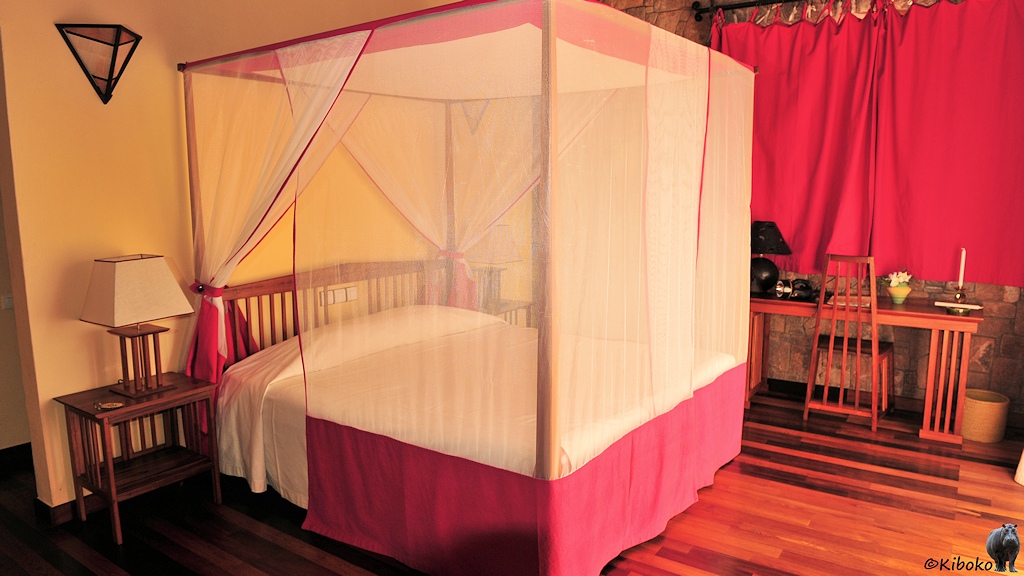 Das Bild zeigt eine Innenansicht von einem Zimmer. In der Mitte steht ein Doppelbett mit einem Holzgestell über das ein Mosikonetz gespannt ist. Im Hintergrund stehen ein schmaler Tisch mit einem Holzstuhl vor einem roten Vorhang.
