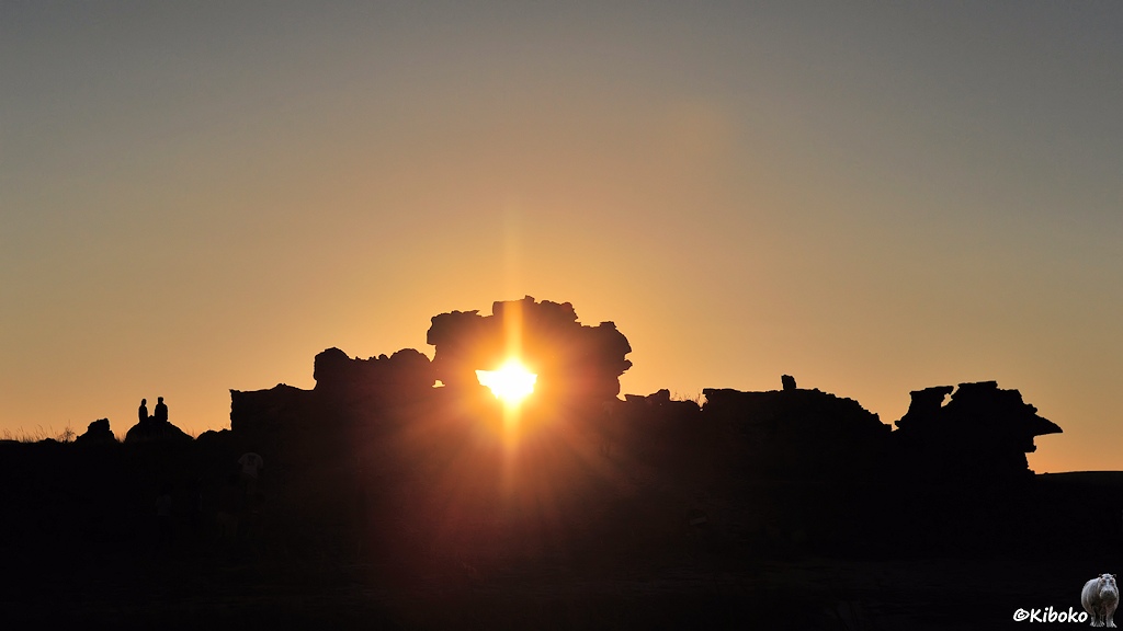 Das Bild zeigt eine Felsformation im Gegenlicht. In der Mitte ist eine größere Öffnung, durch die die untergehende Sonne scheint.