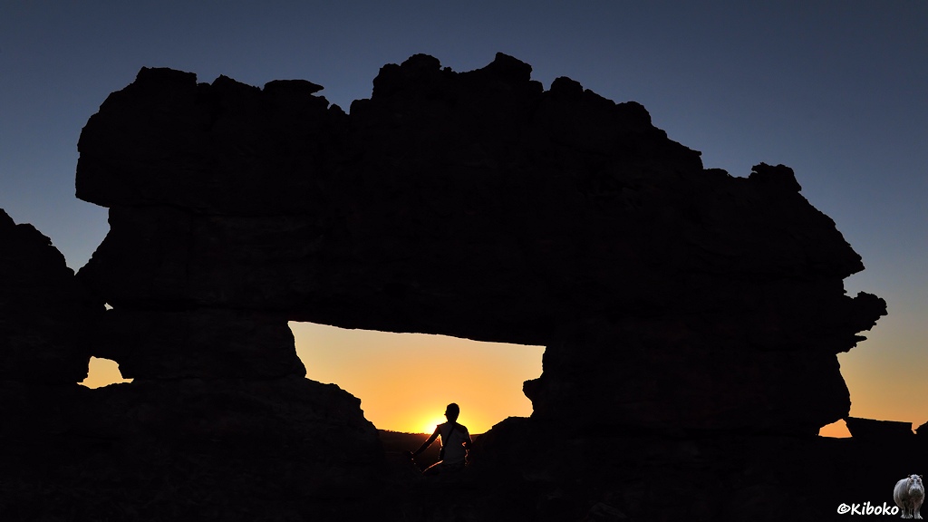 Das Bild zeigt eine natürliche Brücke im Gegenlicht. Eine Frau sitzt genau in der Öffnung vor der untergehenden Sonne. Der Himmel ist dunkelblau und wird nach unten orange. Der Felsen ist als Scherenschnitt schwarz.