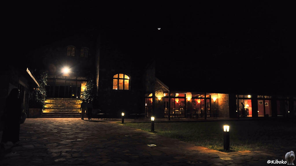 Das Bild zeigt eine Nachtaufnahme von den Weg zum Restaurant aus Natursteinplatten. Durch die Fenster des dahinterliegenden Gebäudes sind Tische und Stühle zu sehen.