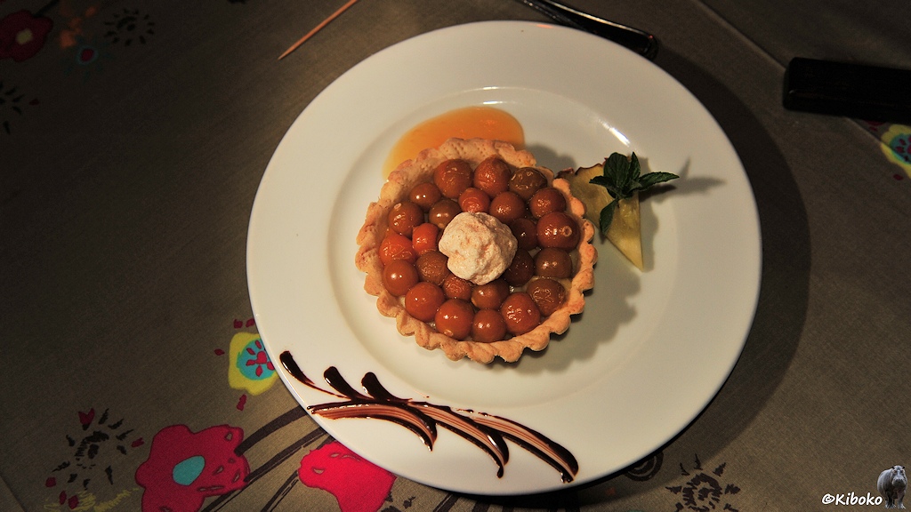 Das Bild zeigt einen weißen Teller mit einem kleinen runden Kuchen mit rotbraunen, kirschgroßen Früchen. Obendrauf ist ein Schlag Sahne.