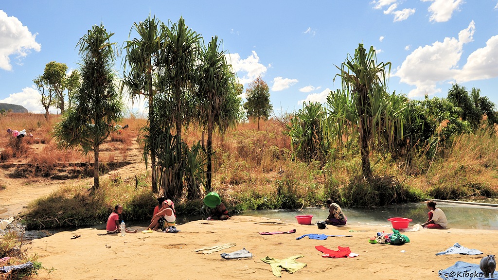 Das Bild zeigt sechs Frauen die am Straßenrand an und in einem kleinen Fluss sitzen und Wäsche waschen. Wäschestücke liegen zum Trocknen auf dem Weg. Eine Frau badet oben ohne und hält sich eine grüne Plastikschüssel vor das Gesicht. Im Hintergrund stehen vereinzelte Bäume.