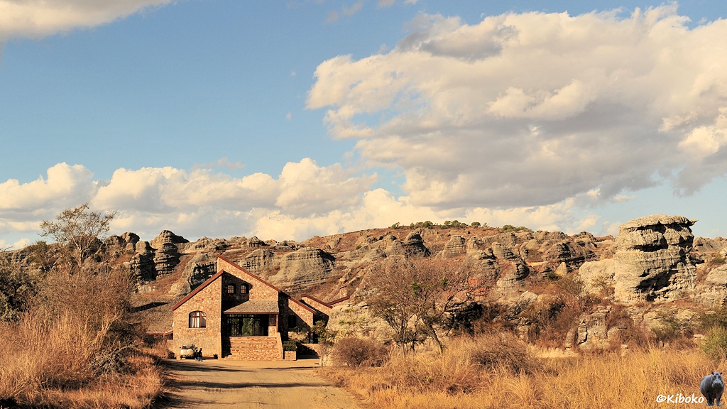 Das Bild zeigt einen beigen Sandweg der auf ein Haus aus braunem Naturstein führt. Im Hintergrund ist eine Felslandschaft aus Hellgrauen Felsen.