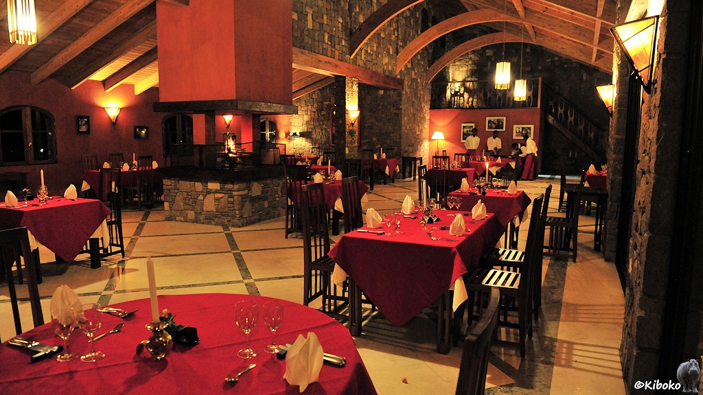 Das Bild zeigt einen beleuchteten Innenarum mit gedeckten Tischen mit roten Tischdecken. Im Hintergrund stehen drei Kellner im weißen Jacket.