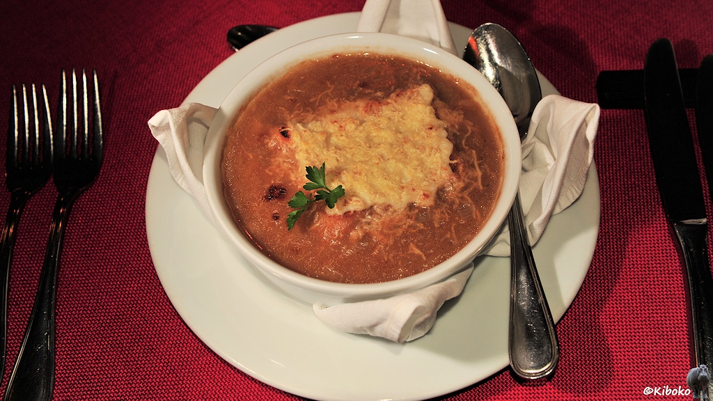Das Bild zeigt eine gefüllte Suppenterrine auf einem runden Teller. Darin ist ein braune Brühe mit einem schwimmenden viereckigen Käse.