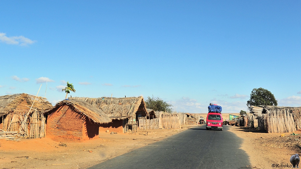 Das Bild zeigt eine gerade Teerstraße durch ein Dorf. Voraus fähr ein roter Kleinbus mit einer hohen Dachladung unter einer blauen Plane. Auf beiden Seiten stehen einfache Häuser mit Schifldach und rotbraunen Lehmwänden. Teilweise ist der Lehm abgebröckelt und die Holzstruktur der Hütte sichtbar.