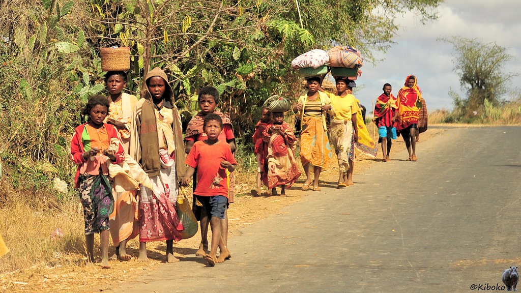 Das Bild zeigt eine Gruppe Frauen und Kinder in bunten Kleidern an einer Teerstraße entgegenkommen. Die Frauen tragen Körbe und Schüsseln mit Wäsche auf dem Kopf. Fast alle sind barfuss.