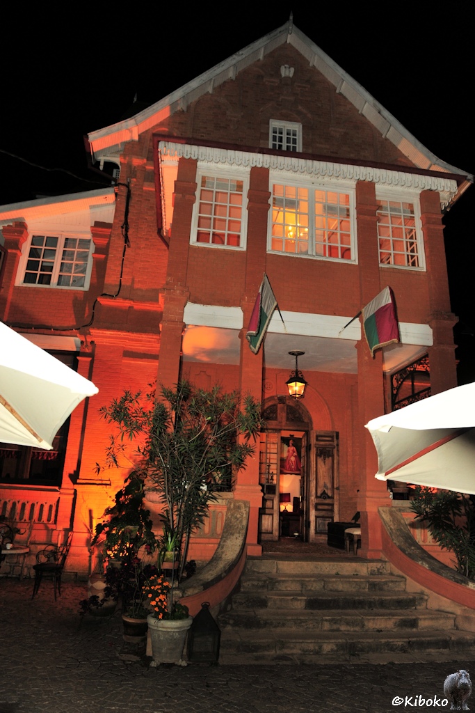 Das Bild zeigt eine Hochformataufnahme eines zweitöckigen, rotbraunen Gebäudes bei Nacht. Eine Treppe führt zur zentralen, geöffneten Eingangstür. Zwei Madagassiche Fahnen wehen über dem Eingang.